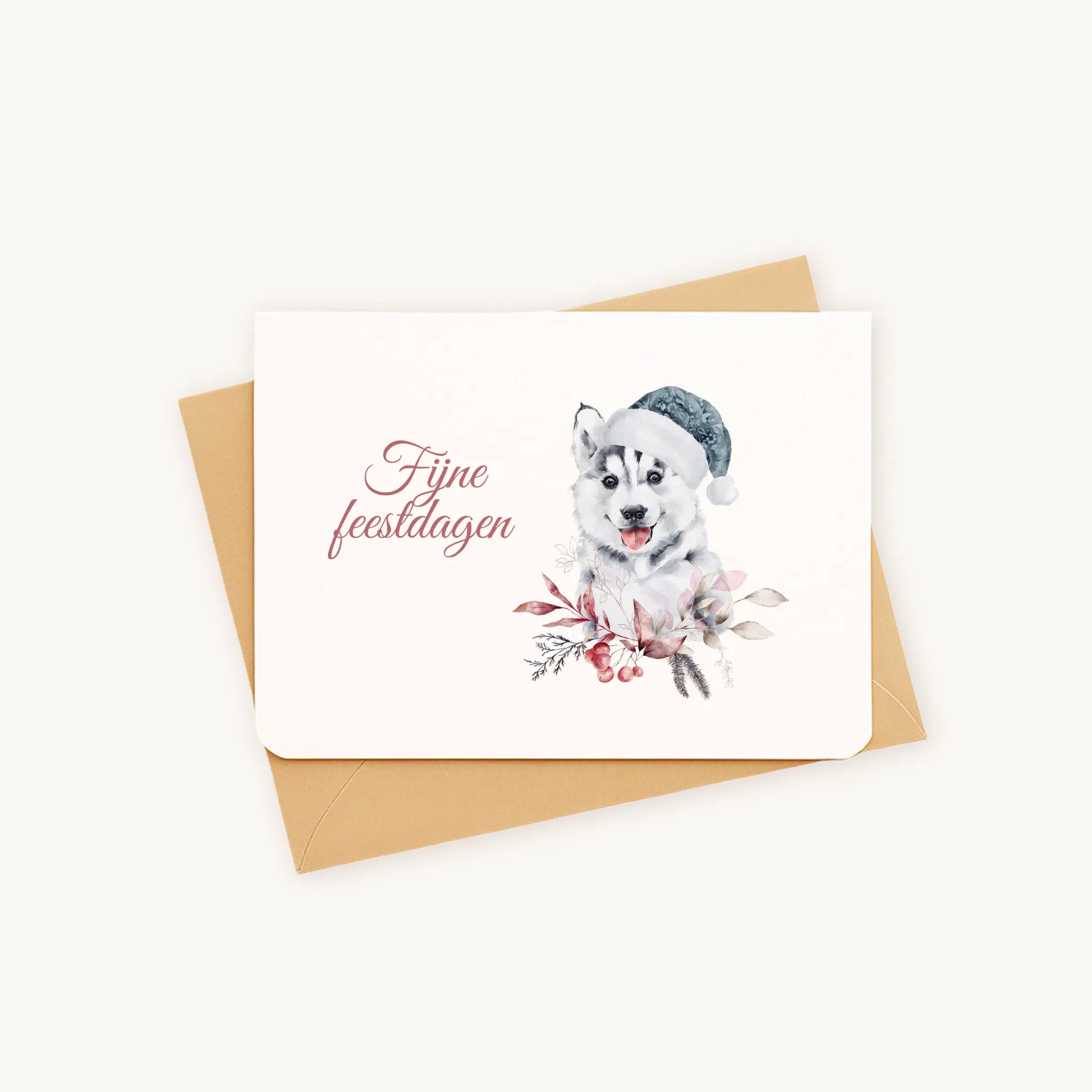 Liggende kerstkaart met aquarel husky-pup met blauwe kerstmanmuts en winterse bladeren en bessen, sierlijke tekst ‘fijne feestdagen’, gepresenteerd op kraftenvelop op lichte achtergrond.