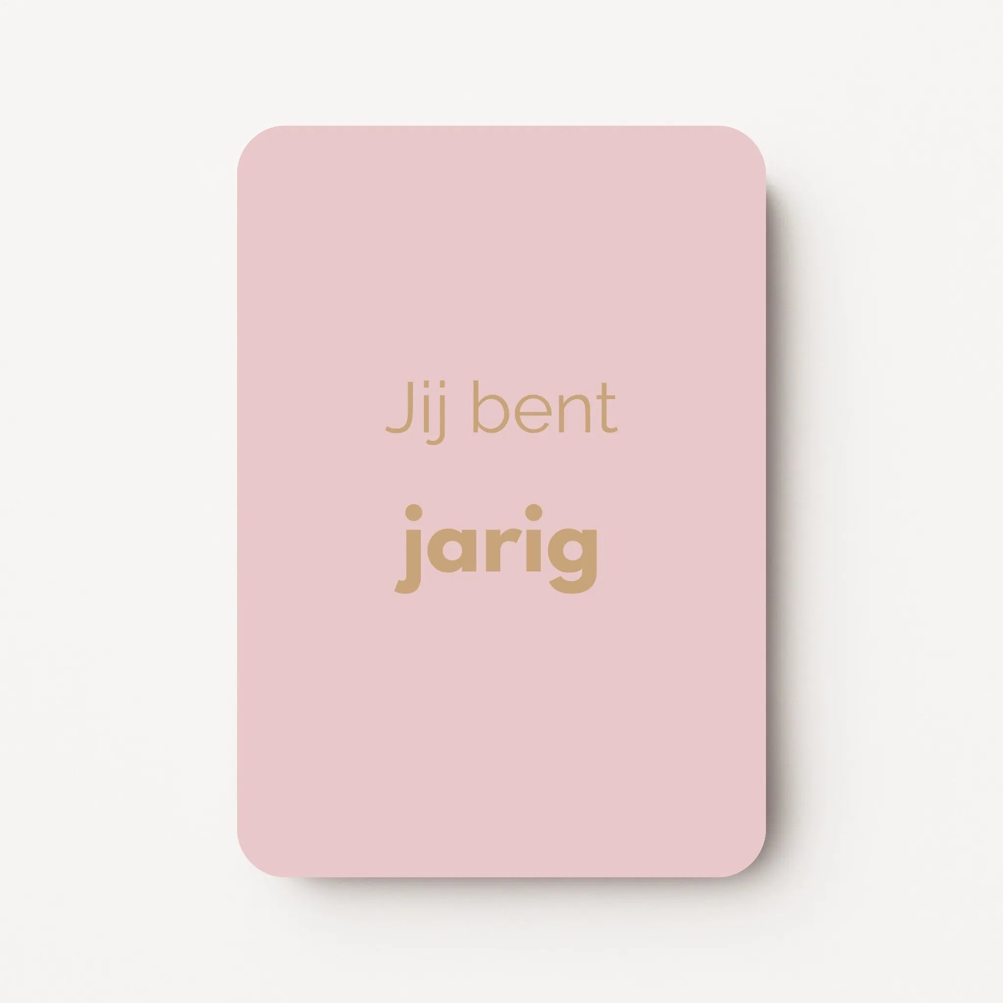 Staand minikaartje met goudbeige tekst ‘Jij bent jarig’, minimalistisch ontwerp op zachte lichtroze achtergrond, flatlay op witte ondergrond.