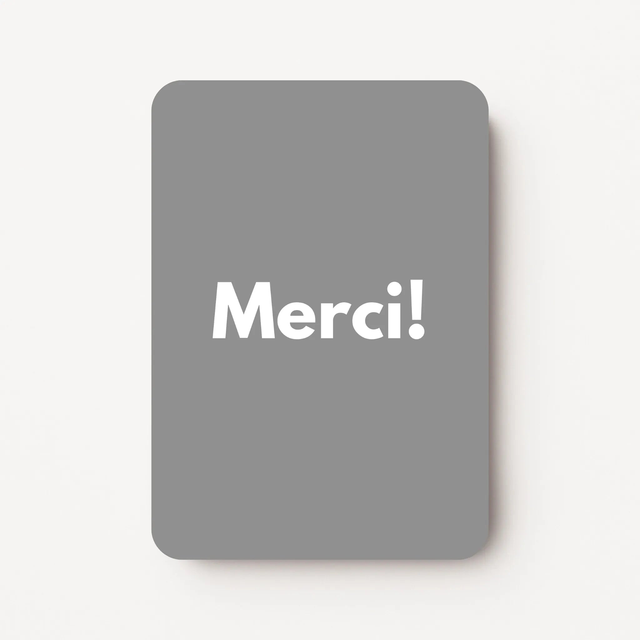 Staand minikaartje met witte tekst ‘Merci!’ op middengrijze achtergrond, minimalistisch ontwerp, flatlay op witte ondergrond.