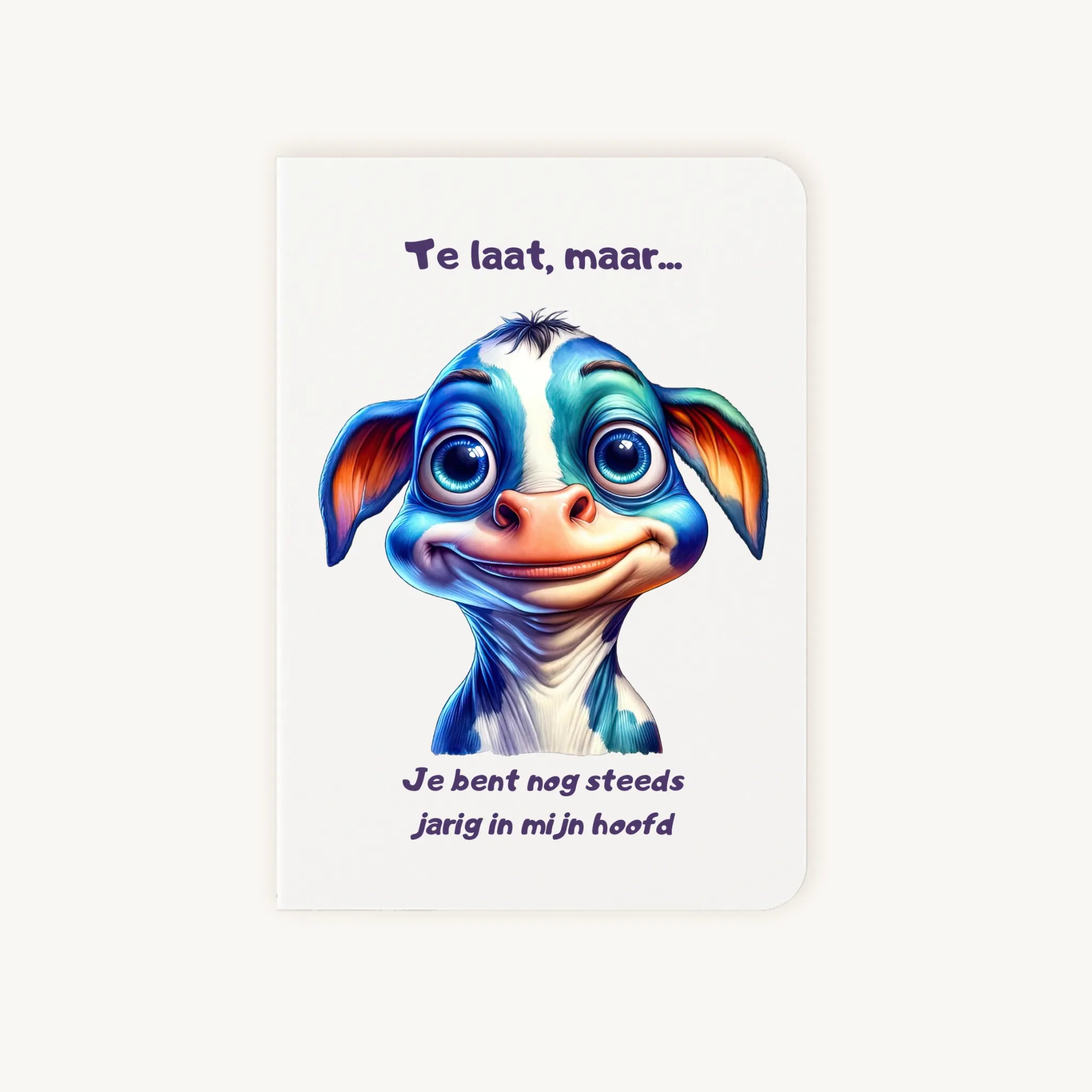 Staande dubbelgevouwen verjaardagskaart met schattige cartoonkoe met grote blauwe ogen en lieve glimlach, met humoristische tekst Te laat maar je bent nog steeds jarig in mijn hoofd, flatlay op lichte achtergrond.