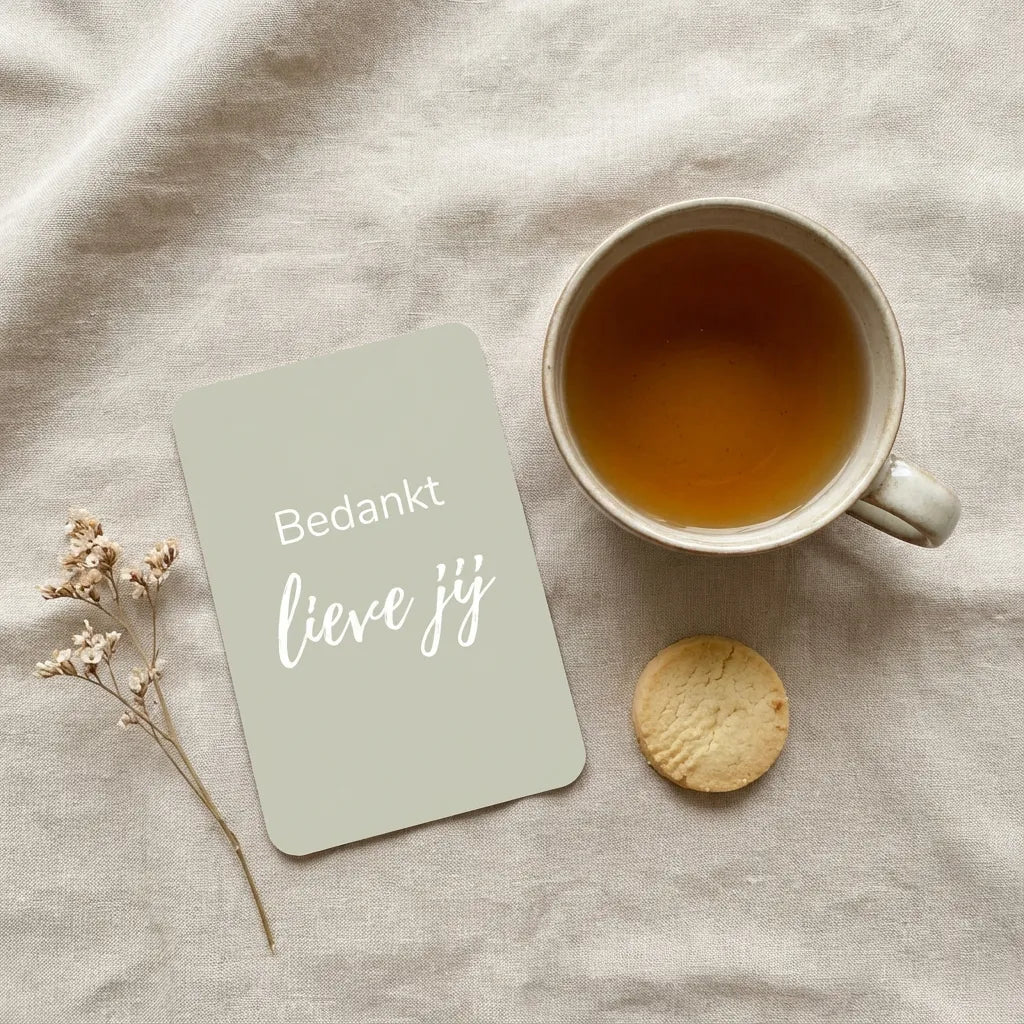 Mini bedankkaart met afgeronde hoeken naast kopje thee, koekje en gedroogd bloemetje op linnen ondergrond
