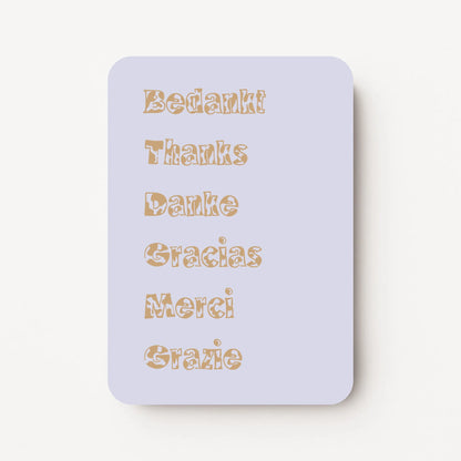 Staande bedankkaart met de woorden ‘Bedankt, Thanks, Danke, Gracias, Merci, Grazie’ onder elkaar in sierlijke letters, goudbruine tekst op lavendel achtergrond, flatlay op witte ondergrond.