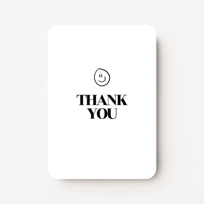 Staande bedankkaart met tekst ‘THANK YOU’ in zwarte letters met een zwarte smiley erboven, flatlay op witte achtergrond.