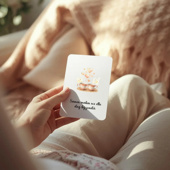 Close-up van een hand die een witte affirmatiekaart in A7-portretformaat met afgeronde hoeken vasthoudt – zachte natuurlijke belichting, warme plaid of kussen op de achtergrond, rustgevende huiselijke sfeer in beige en zachte rozetinten, focus op de kaart en het moment.