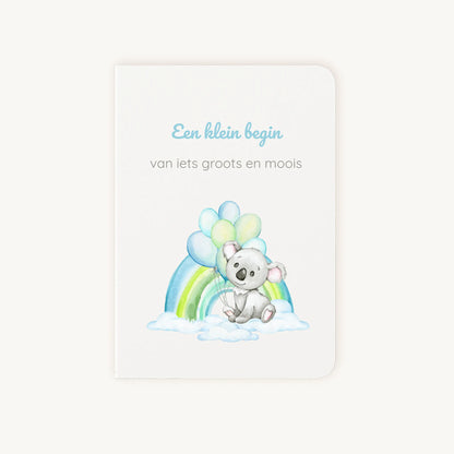 Staande dubbelgevouwen felicitatiekaart geboorte jongen met zachte pastelkleuren, koala met ballonnen en regenboog op wolkenachtergrond, met de tekst "Een klein begin van iets groots en moois", flatlay op lichte achtergrond.