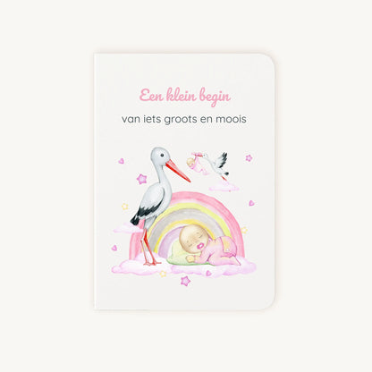 Staande dubbelgevouwen felicitatiekaart geboorte meisje met zachte pastelkleuren, ooievaar met baby onder regenboog, met de tekst "Een klein begin van iets groots en moois", flatlay op lichte achtergrond.