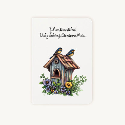 Staande dubbelgevouwen felicitatiekaart nieuwe woning met afgeronde hoekjes en sfeervolle aquarelillustratie van een houten vogelhuisje met bloemen en twee vogeltjes, met de tekst Tijd om te nestelen, flatlay op lichte achtergrond.