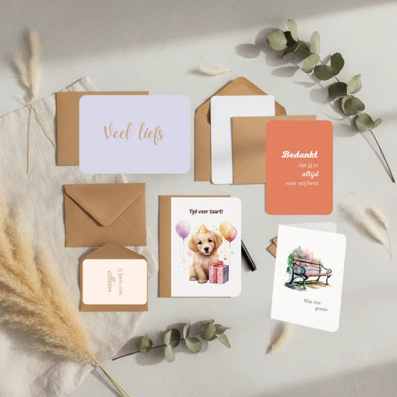 Flatlay van zes wenskaarten – een mix van dubbelgevouwen, enkele en minikaarten met afgeronde hoeken op een lichte neutrale ondergrond. Enkele kaarten liggen deels in of naast kraftenveloppen, met subtiele natuurlijke accenten zoals gedroogde eucalyptus en linnenstof. Rustige, tijdloze sfeer in beige- en grijstinten met zachte natuurlijke belichting.