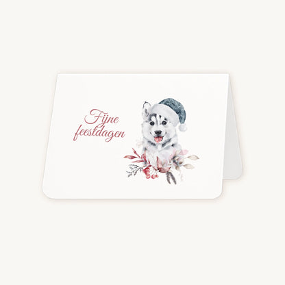 Half geopende liggende kerstkaart met aquarel husky-pup met blauwe kerstmanmuts en winterse bladeren en bessen, sierlijke tekst ‘fijne feestdagen’, op lichte achtergrond.