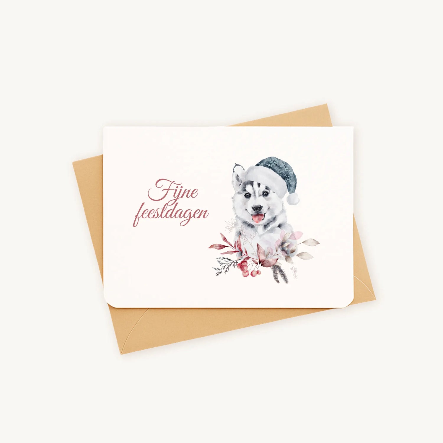 Liggende kerstkaart met aquarel husky-pup met blauwe kerstmanmuts en winterse bladeren en bessen, sierlijke tekst ‘fijne feestdagen’, gepresenteerd op kraftenvelop op lichte achtergrond.