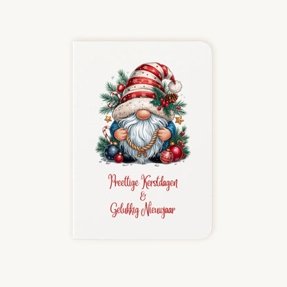 Staande dubbelgevouwen kerstkaart met aquarel kerstkabouter met rood-wit gestreepte muts en sierlijke tekst ‘Prettige Kerstdagen &amp; Gelukkig Nieuwjaar’, flatlay op lichte achtergrond.
