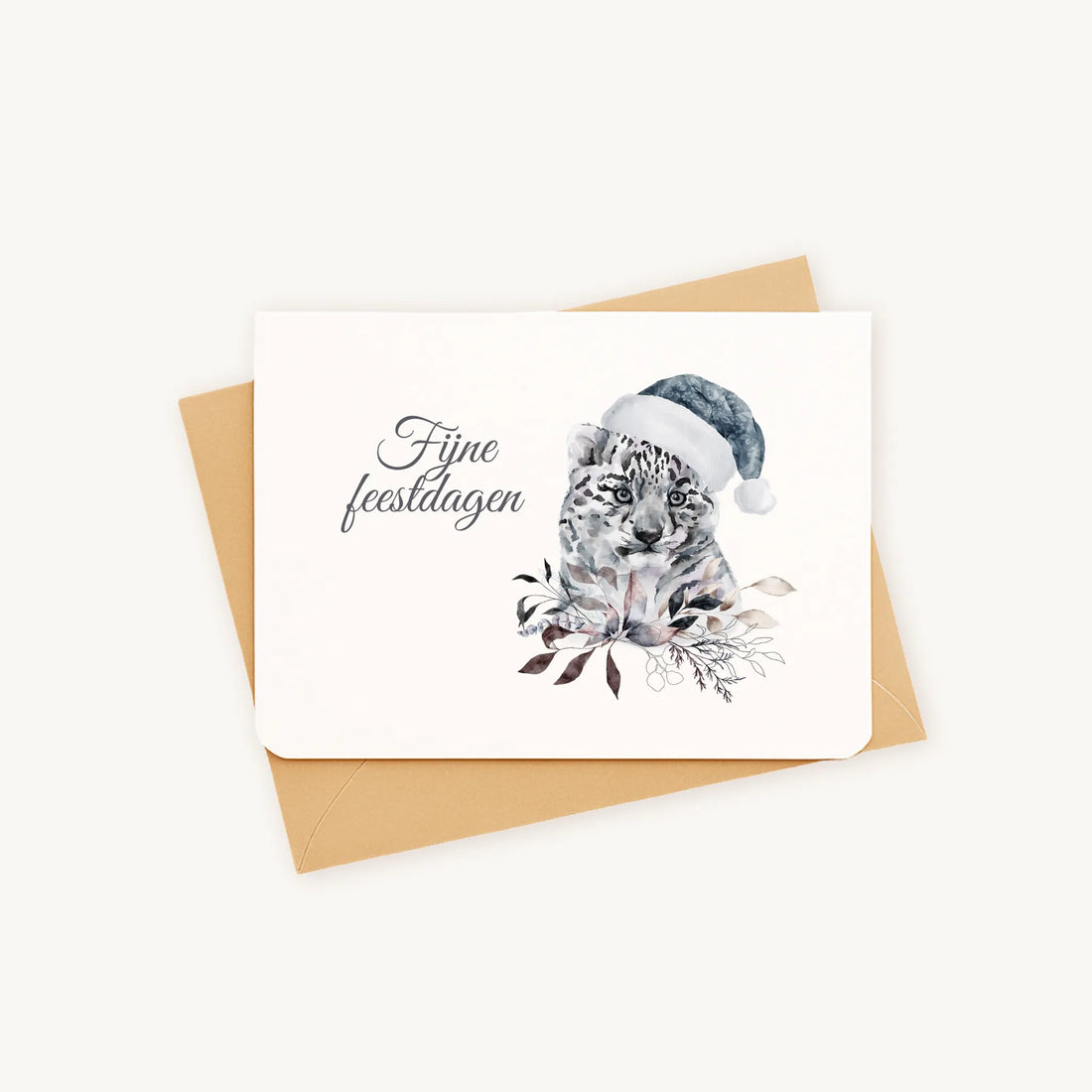 Liggende kerstkaart met aquarel sneeuwluipaard met blauwe kerstmanmuts en wintertakjes en bladeren, sierlijke tekst ‘fijne feestdagen’, op kraftenvelop op lichte achtergrond.