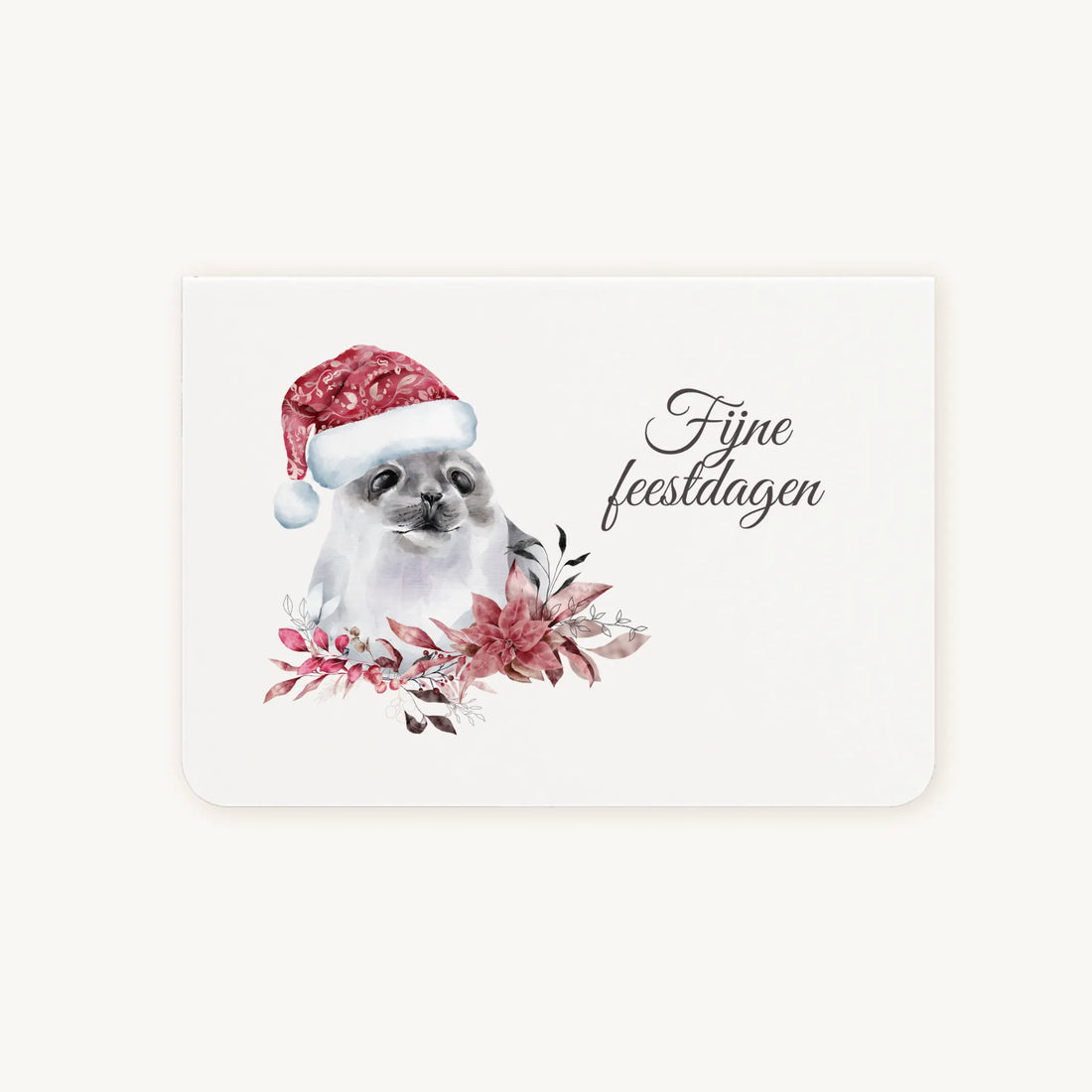 Liggende dubbelgevouwen kerstkaart met aquarel zeehond met kerstmanmuts, omringd door winterse bloemen en blaadjes, sierlijke tekst ‘fijne feestdagen’, flatlay op lichte achtergrond.