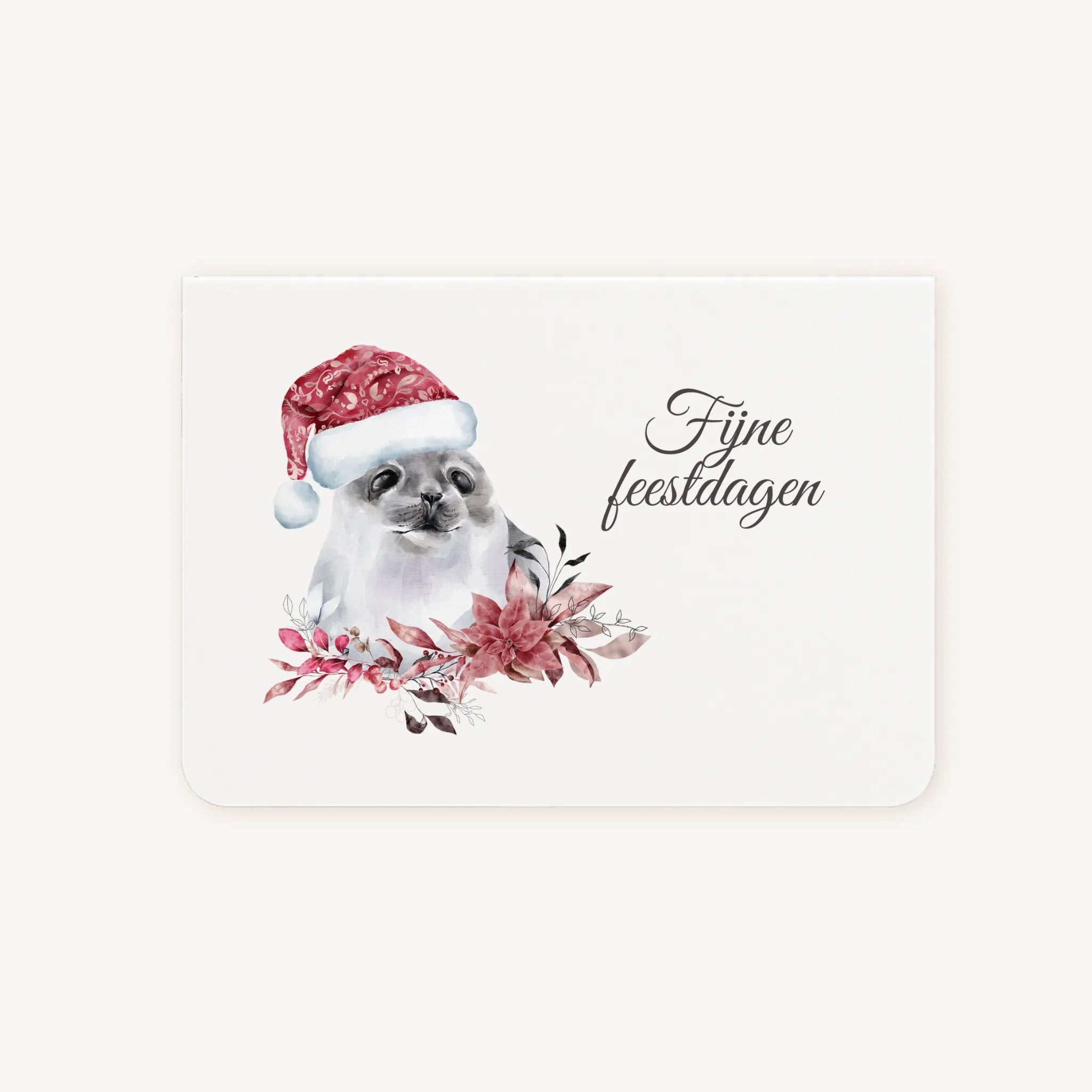 Liggende dubbelgevouwen kerstkaart met aquarel zeehond met kerstmanmuts, omringd door winterse bloemen en blaadjes, sierlijke tekst ‘fijne feestdagen’, flatlay op lichte achtergrond.