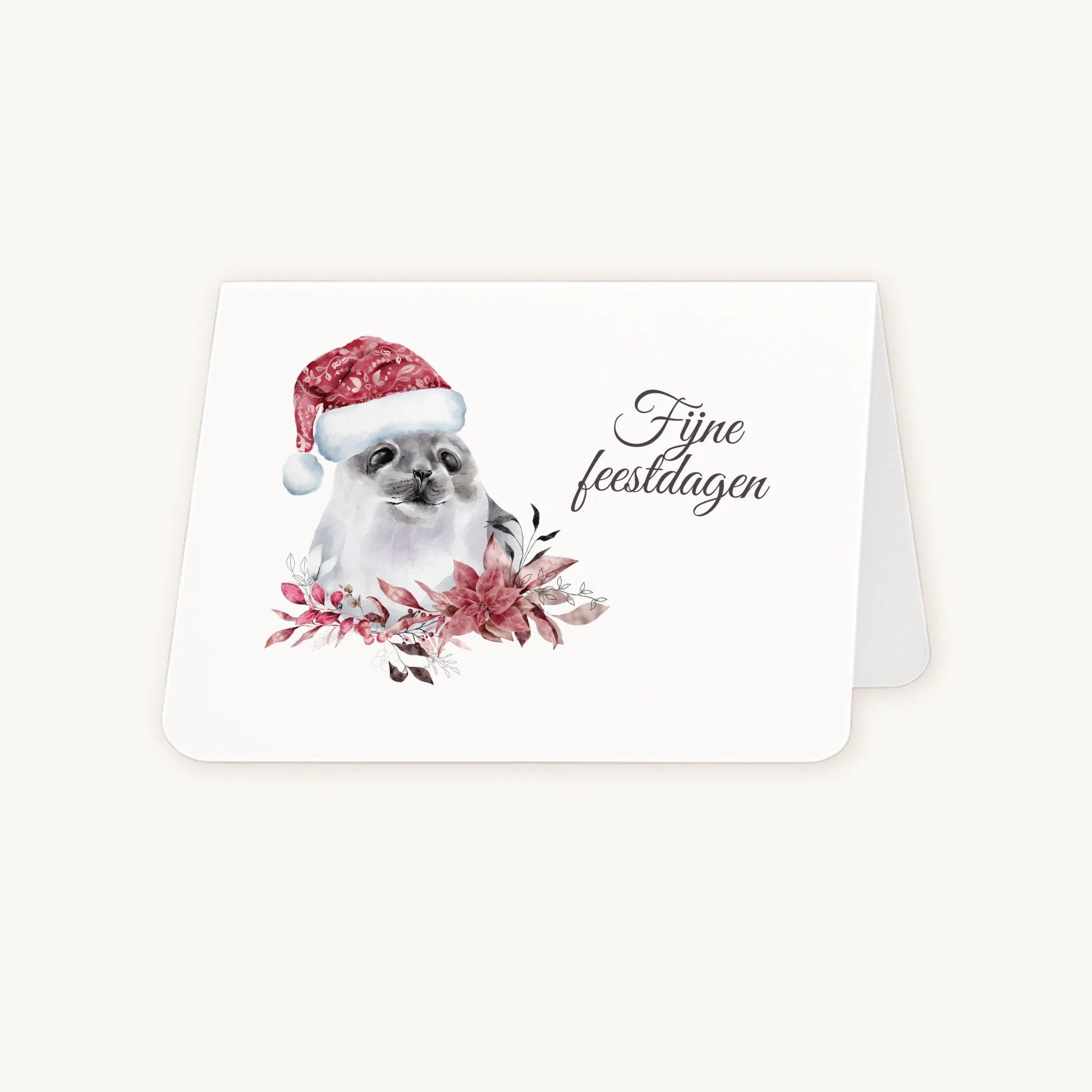 Half geopende liggende kerstkaart met aquarel zeehond met kerstmanmuts en winterse bloemen en blaadjes, sierlijke tekst ‘fijne feestdagen’, op lichte achtergrond.