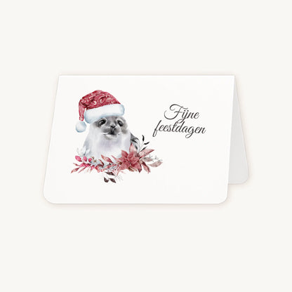 Half geopende liggende kerstkaart met aquarel zeehond met kerstmanmuts en winterse bloemen en blaadjes, sierlijke tekst ‘fijne feestdagen’, op lichte achtergrond.