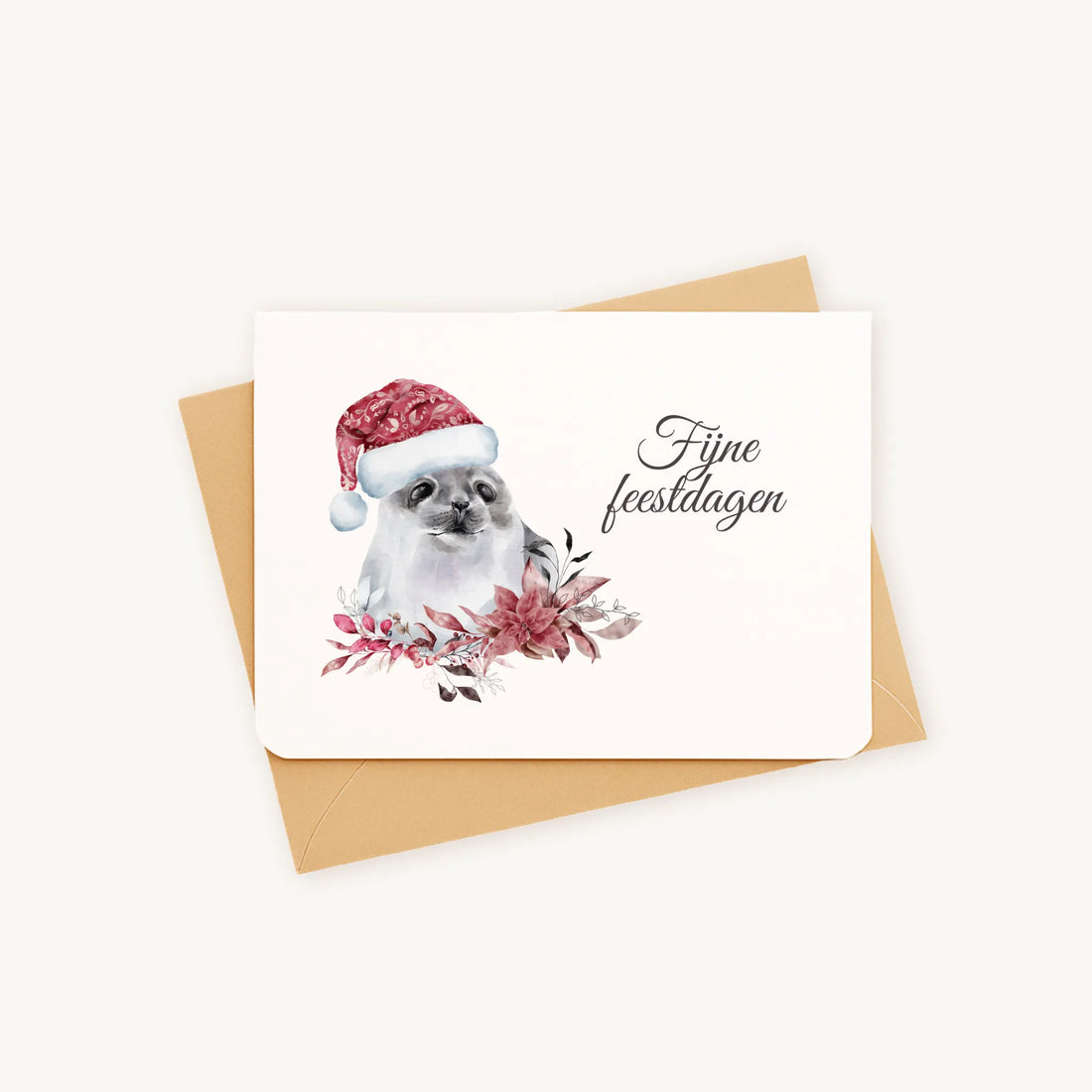 Liggende kerstkaart met aquarel zeehond met kerstmanmuts en winterse bloemen en blaadjes, sierlijke tekst ‘fijne feestdagen’, op kraftenvelop op lichte achtergrond.