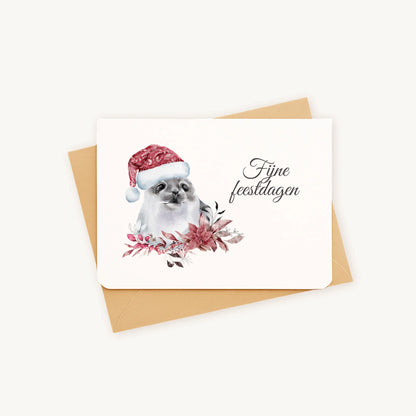 Liggende kerstkaart met aquarel zeehond met kerstmanmuts en winterse bloemen en blaadjes, sierlijke tekst ‘fijne feestdagen’, op kraftenvelop op lichte achtergrond.