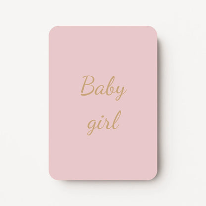 Staand minikaartje met tekst ‘Baby girl’ in goudbeige letters, minimalistisch ontwerp op zachte warmgrijze achtergrond met een vleugje roze, flatlay op witte ondergrond.