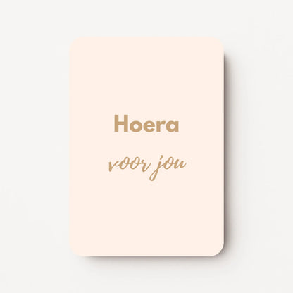 Staand minikaartje met goudbeige tekst ‘Hoera voor jou’, minimalistisch ontwerp op zachte roomkleurige achtergrond, flatlay op witte ondergrond.