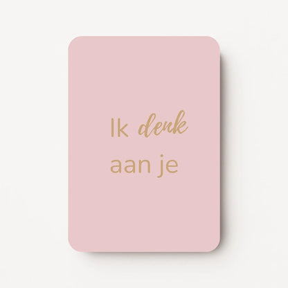 Staand minikaartje met goudbeige tekst ‘Ik denk aan je’, minimalistisch ontwerp op een zachte lichtroze achtergrond, flatlay op witte ondergrond.