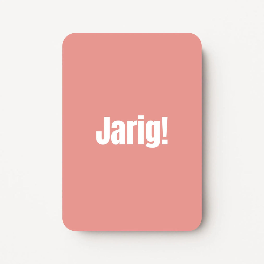 Staand minikaartje met witte tekst ‘Jarig!’, minimalistisch ontwerp op zachte roodroze achtergrond, flatlay op witte ondergrond.