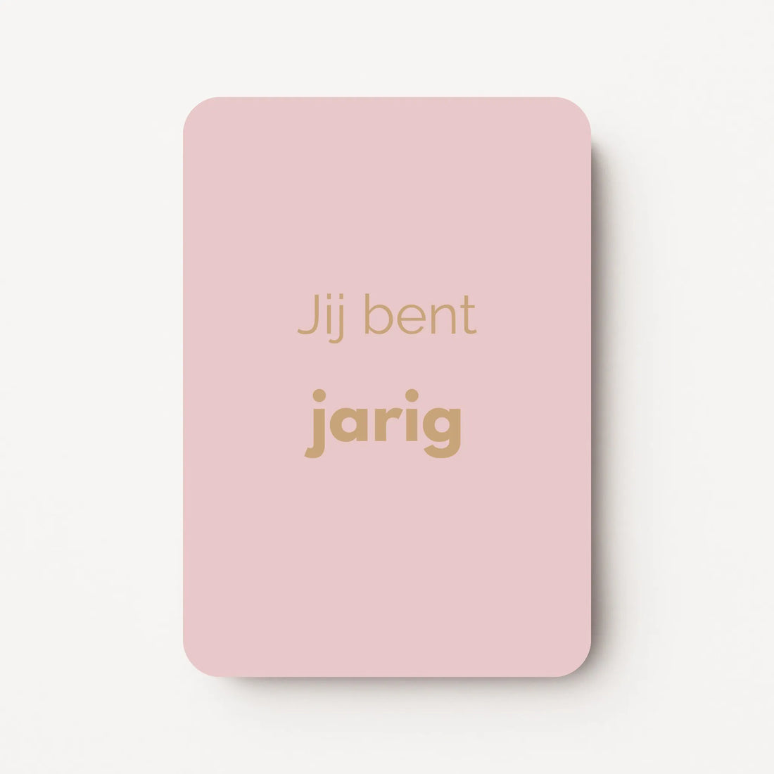 Staand minikaartje met goudbeige tekst ‘Jij bent jarig’, minimalistisch ontwerp op zachte lichtroze achtergrond, flatlay op witte ondergrond.
