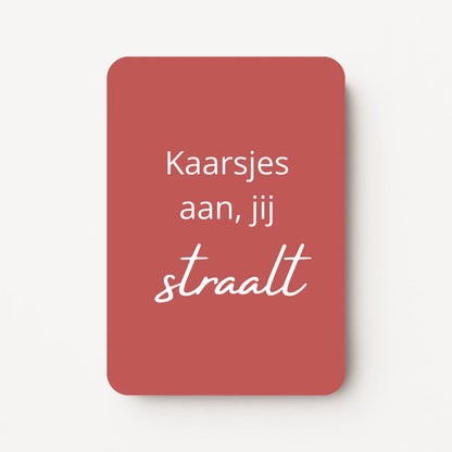 Staand minikaartje met witte tekst ‘Kaarsjes aan, jij straalt’, minimalistisch ontwerp op warme roodbruine achtergrond, flatlay op witte ondergrond.
