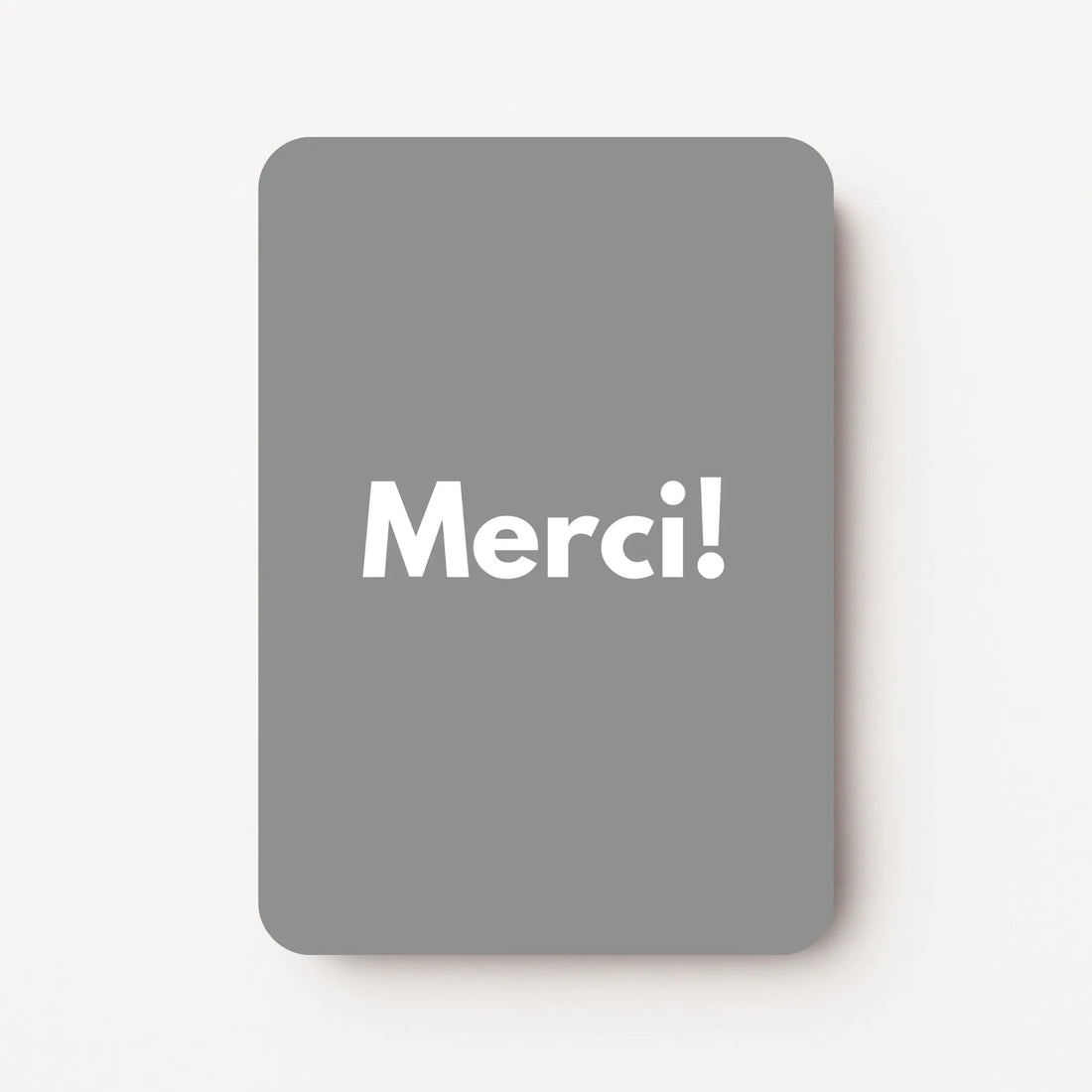 Staand minikaartje met witte tekst ‘Merci!’ op middengrijze achtergrond, minimalistisch ontwerp, flatlay op witte ondergrond.