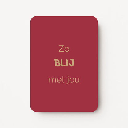 Staand minikaartje met tekst ‘Zo BLIJ met jou’ in goudbeige letters, minimalistisch ontwerp op diepe roodoranje achtergrond, flatlay op witte ondergrond.