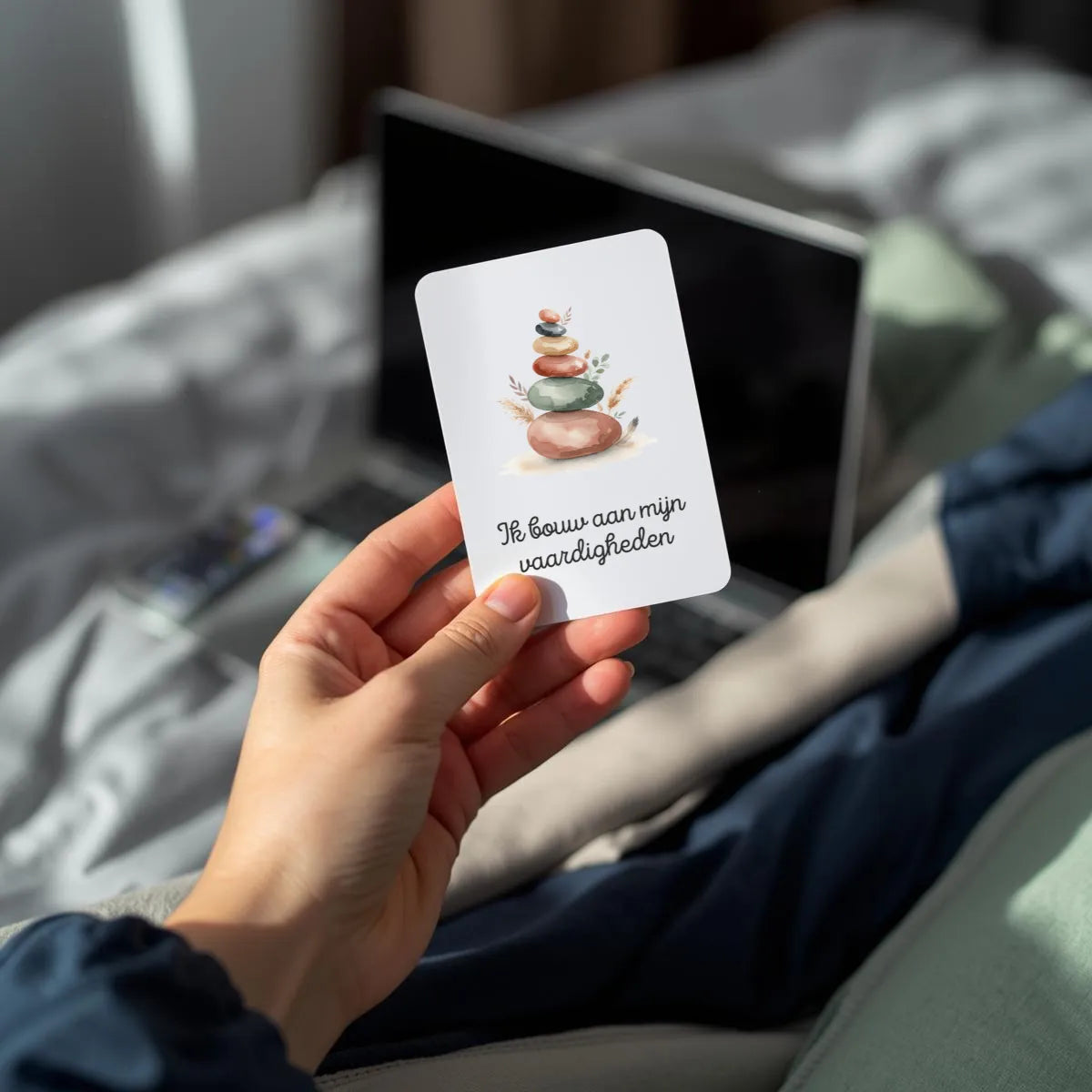 Tienerhand (13–17 jaar) houdt een lege witte A7-kaart met afgeronde hoeken rechtop vast – zachte natuurlijke belichting, laptop of boek vaag op de achtergrond, moderne rustige sfeer met grijs, navyblauw en zachtgroen, focus op de blanco kaart en het natuurlijke moment.