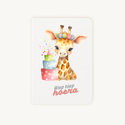 Staande dubbelgevouwen verjaardagskaart met schattige giraf met feesthoedje en kleurrijke verjaardagstaart, met tekst Hiep hiep hoera, flatlay op lichte achtergrond.