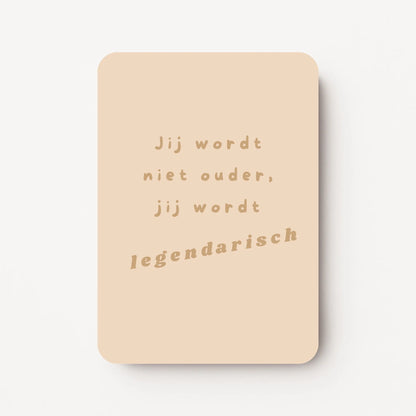 Staande verjaardagskaart voor mannen met goudbeige tekst ‘Jij wordt niet ouder, jij wordt legendarisch’ op een warme beige achtergrond, flatlay op witte ondergrond.