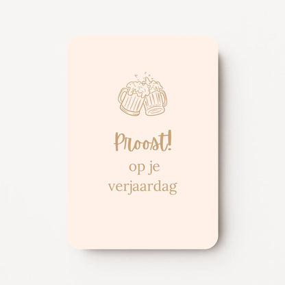 Staande verjaardagskaart voor mannen met goudbeige tekst ‘Proost! op je verjaardag’ en bovenaan een lijntekening van twee proostende bierpullen, op een crèmekleurige achtergrond, flatlay op witte ondergrond.