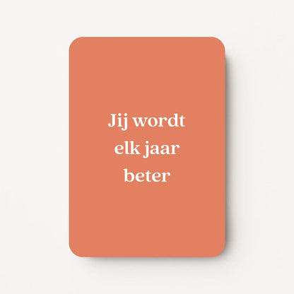 Staande verjaardagskaart voor een man met witte tekst ‘Jij wordt elk jaar beter’ op een warme oranje achtergrond, minimalistisch ontwerp, flatlay op witte ondergrond.