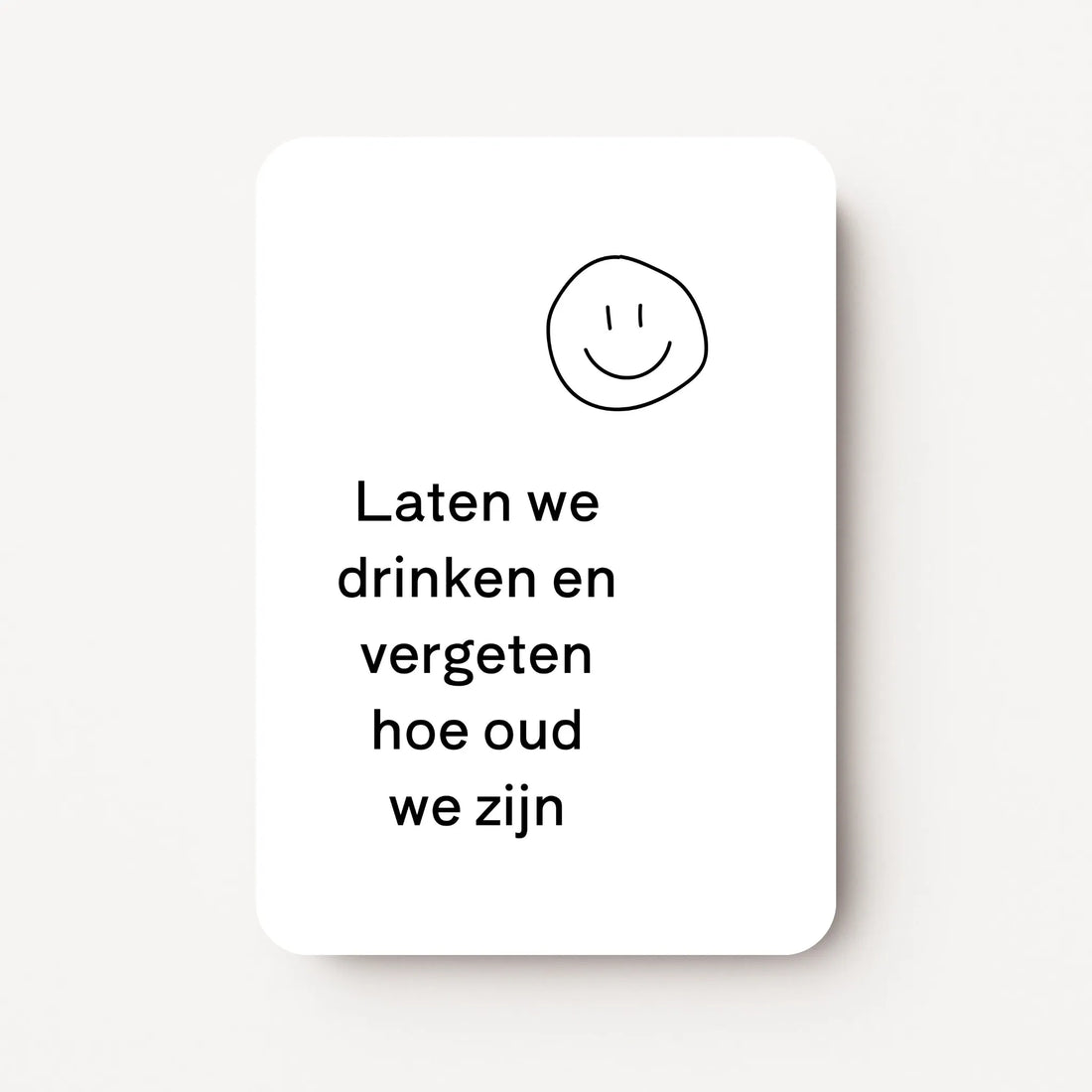 Staande verjaardagskaart voor mannen met zwarte tekst ‘Laten we drinken en vergeten hoe oud we zijn’ en een getekende smiley erboven, minimalistisch ontwerp op witte achtergrond, flatlay op witte ondergrond.