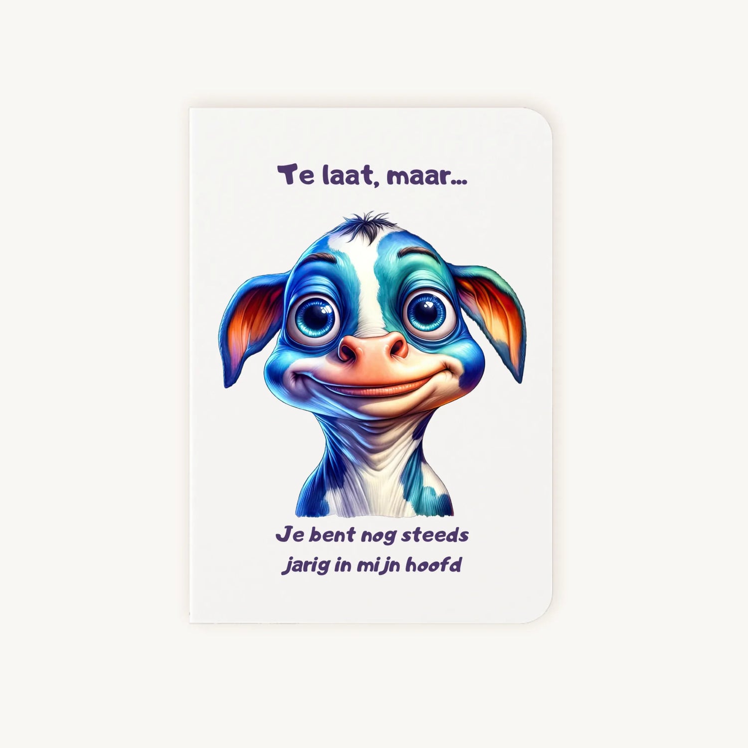 Staande dubbelgevouwen verjaardagskaart met schattige cartoonkoe met grote blauwe ogen en lieve glimlach, met humoristische tekst Te laat maar je bent nog steeds jarig in mijn hoofd, flatlay op lichte achtergrond.