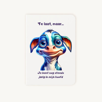 Staande dubbelgevouwen verjaardagskaart met schattige cartoonkoe met grote blauwe ogen en lieve glimlach, met humoristische tekst Te laat maar je bent nog steeds jarig in mijn hoofd, flatlay op lichte achtergrond.