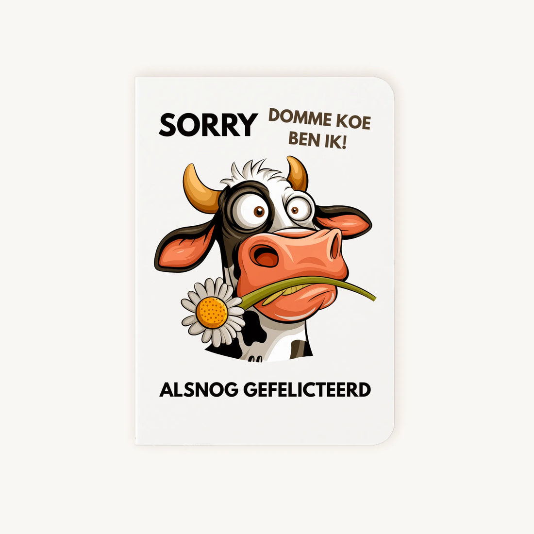 Staande dubbelgevouwen verjaardagskaart met verbaasde koe met madeliefje en tekst ‘SORRY domme koe ben ik – Alsnog gefeliciteerd’, flatlay op lichte achtergrond.