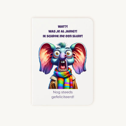 Staande dubbelgevouwen verjaardagskaart met cartoonachtige geschrokken olifant met grote oren en kleurrijke sjaal, met humoristische tekst Wat?! Was je al jarig? Ik schrok me een slurf! Nog steeds gefeliciteerd, flatlay op lichte achtergrond.
