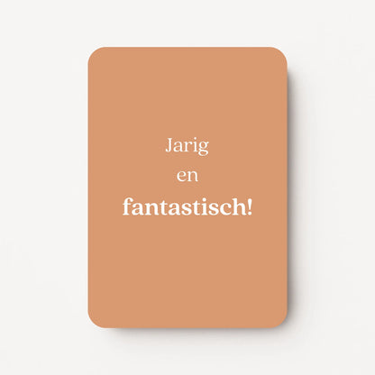 Staande verjaardagskaart voor een vrouw met witte tekst ‘Jarig en fantastisch!’ op een warme zandkleurige achtergrond, flatlay op witte ondergrond.