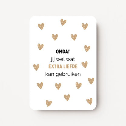 Staande kaart met zwarte tekst ‘Omdat jij wel wat extra liefde kan gebruiken’ waarbij ‘extra liefde’ en kleine hartjes in goudbruine kleur zijn, op witte achtergrond, flatlay op witte ondergrond.