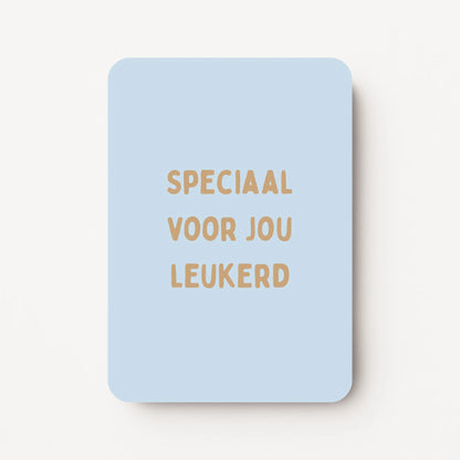 Staande kaart met tekst ‘Speciaal voor jou leukerd’ in goudbruine letters op een zachtblauwe achtergrond, flatlay op witte ondergrond.
